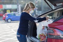 Жінка в масці для обличчя завантажує продукти в машину на парковці — стокове фото