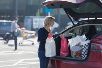 Mujer cargando comestibles en la parte trasera del coche en el estacionamiento soleado - foto de stock