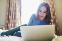 Teenage girl using laptop on bed in sunny bedroom — Stock Photo
