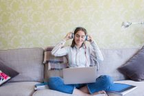 Ragazza sorridente adolescente con le cuffie utilizzando il computer portatile sul divano del soggiorno — Foto stock