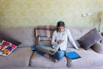 Teenage girl with headphones and laptop using smart phone on sofa — Stock Photo