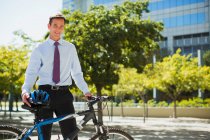 Homme d'affaires souriant avec vélo et casque dans un parc urbain — Photo de stock