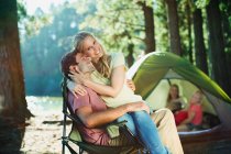 Mujer sonriente sentada en los maridos regazo en el camping en el bosque - foto de stock