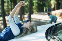 Enthusiastic woman taking self-portrait on hood of car in woods — Stock Photo