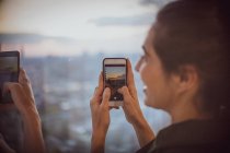 Businesswoman with camera phone photographing sunset at window — Stock Photo