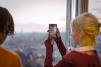 Jovem mulher usando telefone câmera na janela do escritório highrise — Fotografia de Stock