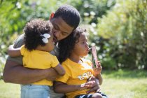 Любящий отец обнимает дочерей на солнечном летнем заднем дворе — стоковое фото