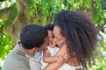 Affectionate parents kissing toddler daughter below tree — Stock Photo
