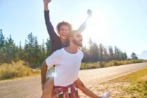 Portrait carefree young couple piggybacking at sunny remote roadside — Stock Photo