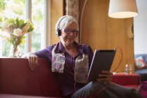 Mulher idosa sorridente com fones de ouvido usando tablet digital no sofá — Fotografia de Stock