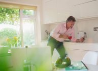 Uomo anziano bere tè e utilizzando smart phone in cucina soleggiata — Foto stock