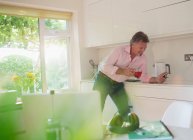 Hombre mayor bebiendo té y utilizando el teléfono inteligente en la cocina soleada - foto de stock