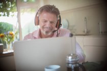 Happy senior man with headphones working at laptop in morning kitchen — Stock Photo
