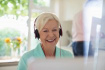 Happy senior woman with headphones using laptop in kitchen — Stock Photo