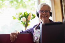 Femme âgée souriante avec écouteurs utilisant une tablette numérique sur le canapé — Photo de stock
