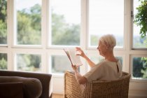 Старша жінка розслабляє і читає книгу в сонячній вітальні — стокове фото