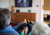 Старша пара тримає руки і дивиться телевізор на дивані у вітальні — стокове фото