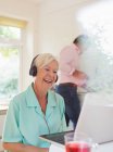 Heureuse femme âgée avec écouteurs vidéo bavardage à l'ordinateur portable dans la cuisine — Photo de stock