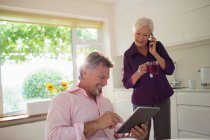 Feliz pareja de ancianos utilizando tableta digital y teléfono inteligente en la cocina - foto de stock