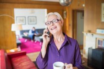 Happy senior woman with coffee talking on smart phone in living room — Stock Photo