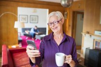 Glückliche Seniorin nutzt Smartphone und trinkt Tee im Wohnzimmer — Stockfoto