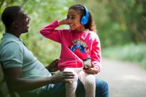 Feliz padre e hija sentados en el banco del parque con auriculares - foto de stock