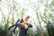 Brincalhão pai despreocupado levantando filha abaixo de árvores no parque ensolarado — Fotografia de Stock