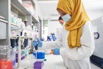 Женщина-ученый в хиджабе и маске для лица с помощью пипетки в лаборатории — стоковое фото