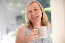 Retrato feliz mujer mayor bebiendo té en la ventana soleada - foto de stock