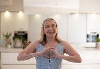 Porträt schöne glückliche Seniorin gestikuliert Herzform in Küche — Stockfoto