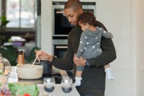 Father holding baby daughter and cooking dinner at kitchen stove — Stock Photo