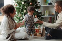 Happy parents and baby daughter opening Christmas gift by tree — Stock Photo