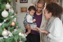 Happy parents helping baby daughter open Christmas gift by tree — Stock Photo