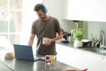 Человек с наушниками работает из дома за ноутбуком на кухне — стоковое фото