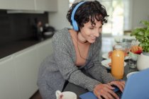 Happy woman with headphones working from home at laptop in kitchen — Stock Photo