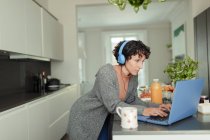 Mujer con auriculares trabajando desde casa en el ordenador portátil en la cocina - foto de stock
