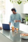 Homme avec écouteurs travaillant de la maison à l'ordinateur portable dans la cuisine du matin — Photo de stock