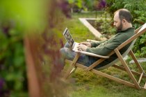 Человек, работающий за ноутбуком в шезлонге в летнем саду — стоковое фото
