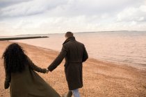 Ласковая пара, держащаяся за руки, гуляющая по пляжу зимнего океана — стоковое фото
