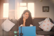 Sorrindo mulher trabalhando em casa falando no telefone inteligente no laptop — Fotografia de Stock