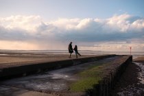 Happy couple in winter coats on sunny ocean beach jetty — Stock Photo