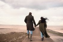 Пара в зимних пальто, держащихся за руки на причале у океана — стоковое фото