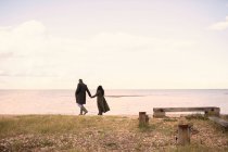 Couple in winter coats holding hands walking on ocean beach — Stock Photo