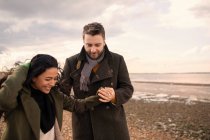 Happy couple in winter coats holding hands walking on beach — Stock Photo