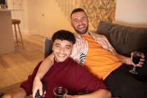 Retrato feliz gay macho pareja bebiendo vino en sofá en casa - foto de stock