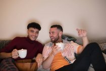 Happy gay male couple drinking hot cocoa watching TV on sofa — Stock Photo