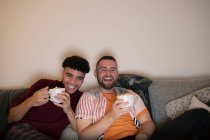 Счастливая гей-пара пьет горячее какао и смотрит телевизор на диване — стоковое фото