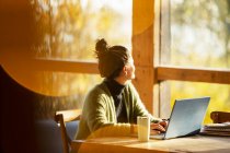 Женщина, работающая за ноутбуком, смотрит в окно в солнечном кафе — стоковое фото