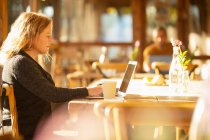Empresária com café trabalhando no laptop no café ensolarado — Fotografia de Stock
