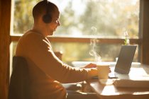 Empresário com fones de ouvido e café quente trabalhando no laptop no café — Fotografia de Stock